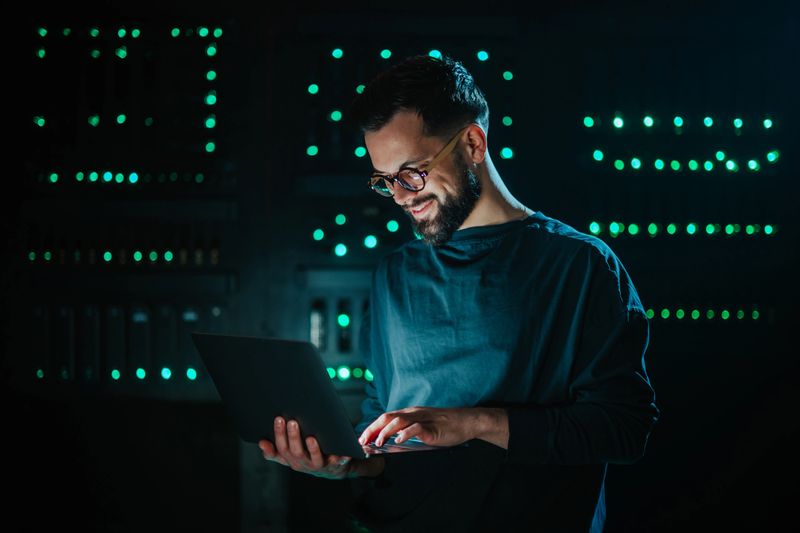 Smiling bearded man working on laptop in dark data center, managing network and database for it security