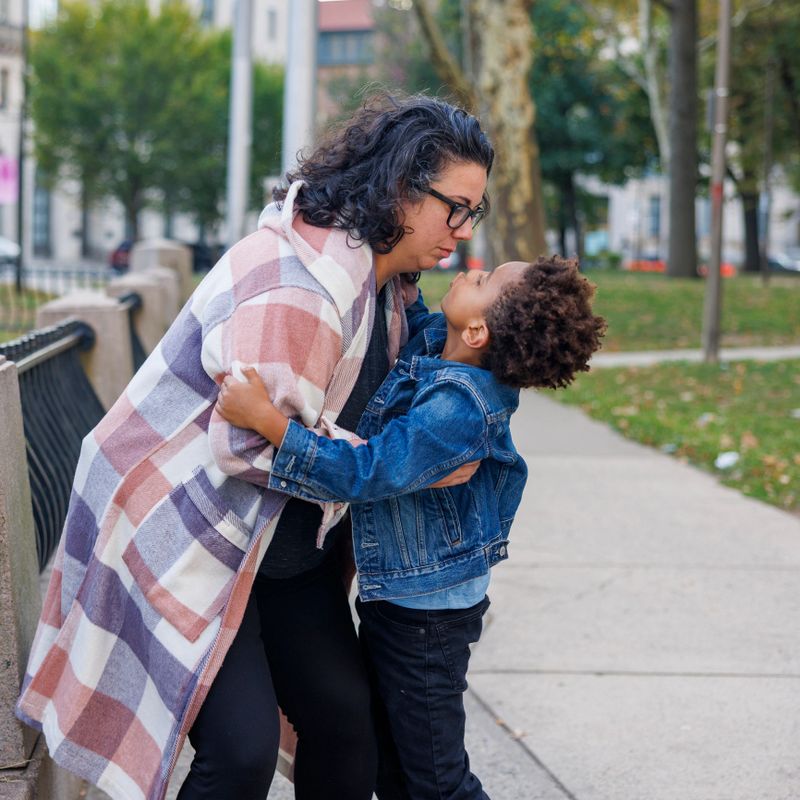 Dark-haired 41 year old Caucasian mother hugging her 7 year old multiracial son on sidewalk. The mother, wearing glasses and a plaid coat, leans down to embrace the boy who looks up at her affectionately. They share a close moment near a fence in Harriet Tubman Square, Newark, New Jersey during an autumn afternoon.