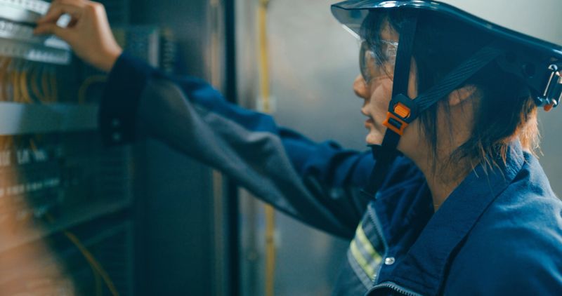 Young Asian female technician in factory, adjusting and examining an electrical control panel to ensure proper function and safety. Robotic Industrial Factory Concept.