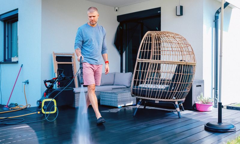 Adult man using a pressure washer to clean a wooden terrace on a modern residential patio. Outdoor home maintenance and seasonal cleaning concept. Real lifestyle scene with contemporary furniture and minimal architecture.