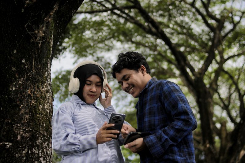 Young Asian woman wearing a hijab and headphones shows a song she is listening to on her smartphone to a young Asian man. Friendship, music sharing, social interaction, and outdoor lifestyle concept