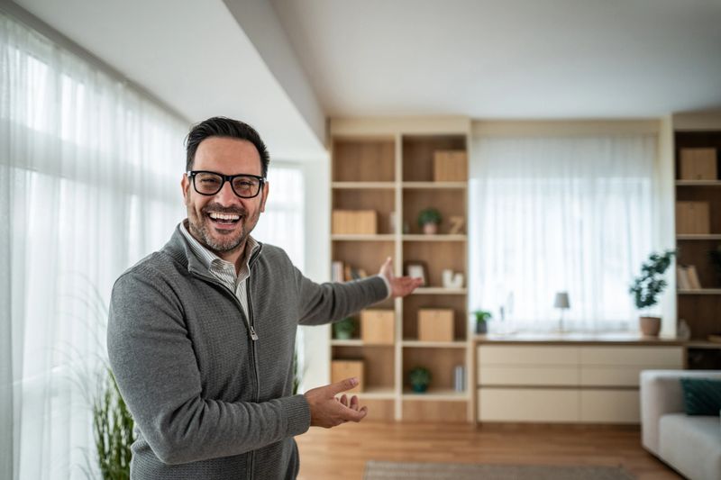 Happy middle-aged man smiling and gesturing with open hands in a bright, minimalist living room, welcoming viewers during an open house or property showing