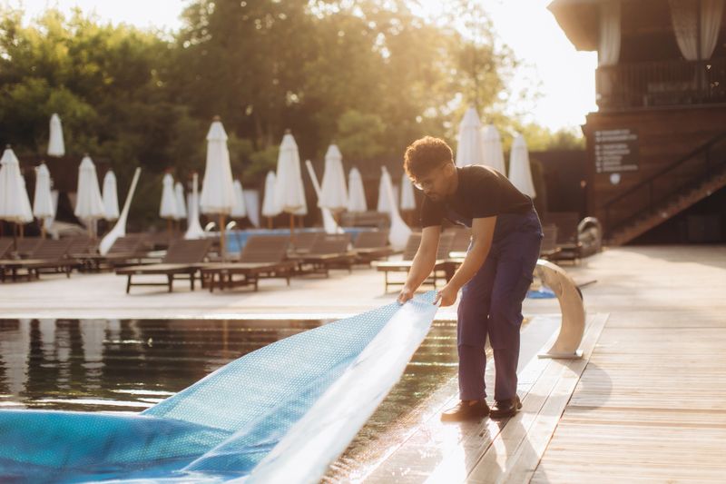 Pool maintenance worker removing a solar cover from a hotel pool in the morning sunshine, preparing for a refreshing summer day