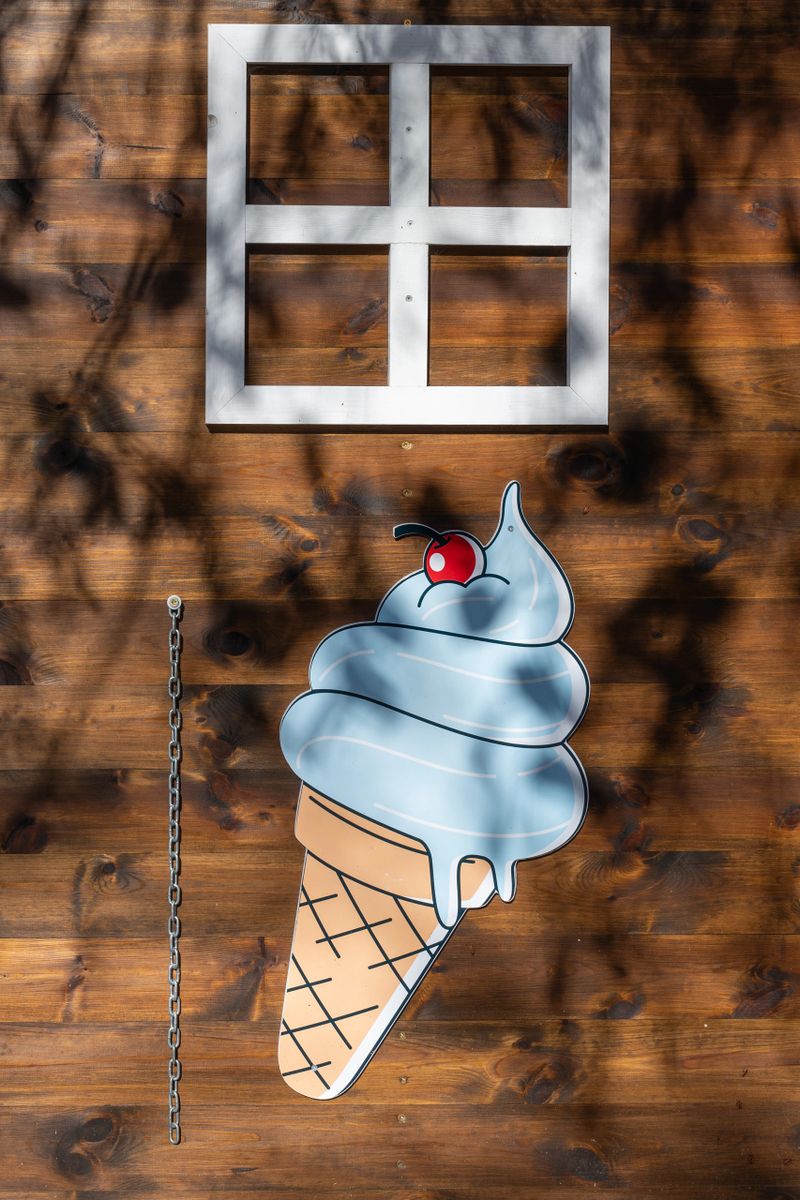 An artisanal outdoor façade composed of stained wooden planks, a square white window frame, and a colorful illustration of a dripping ice cream cone topped with a cherry.