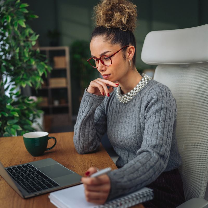 Woman focused on work, typing on a laptop and jotting notes in a spiral notebook at a wooden desk in a modern home office, smiling as she plans and researches remotely