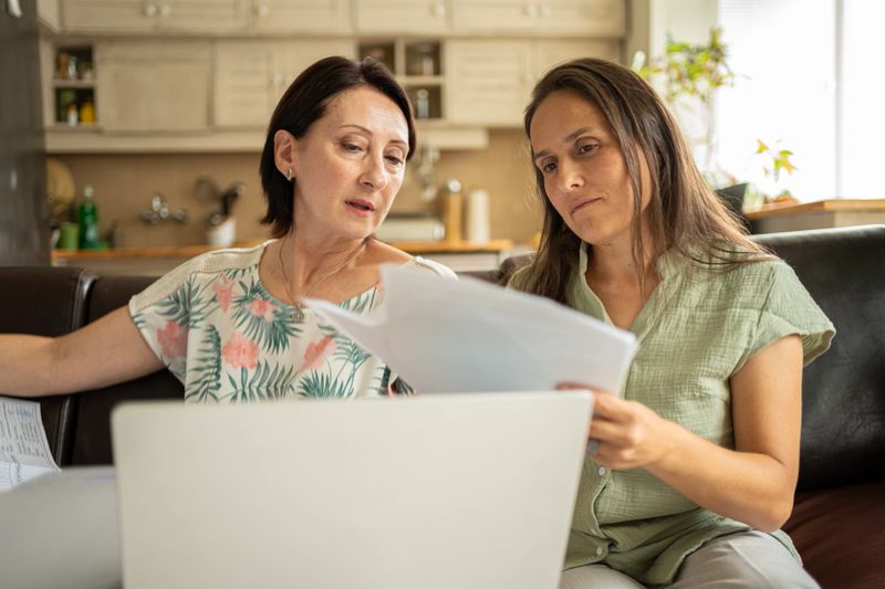 Serious mature women working together, reviewing documents & discussing details of real estate open house event preparation while using laptop for guidance & tips