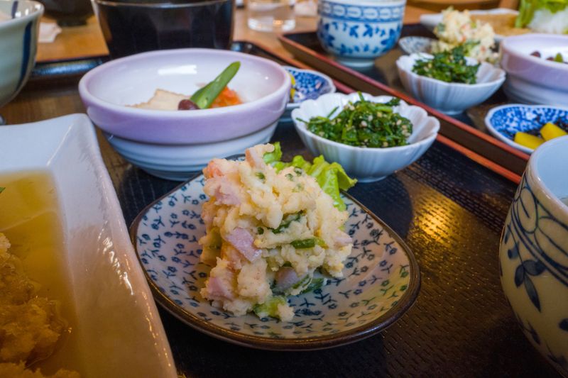 Colorful Japanese potato salad with diced ham and greens is served in a patterned dish, surrounded by assorted vegetable side dishes and ceramic bowls on a lacquered tray. Warm indoor lighting enhances the textures and inviting presentation.