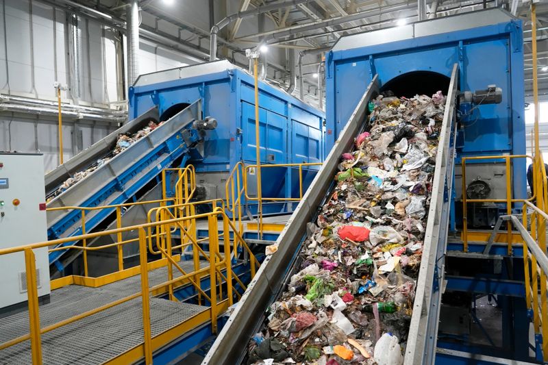 An expansive view inside a modern recycling plant, showcasing two large blue industrial machines with conveyor belts actively moving a substantial volume of unsorted garbage. This image effectively conveys themes of waste management, environmental protection, recycling processes, and the challenges of handling large-scale refuse, suitable for environmental documentaries, industry reports, and sustainability awareness campaigns.