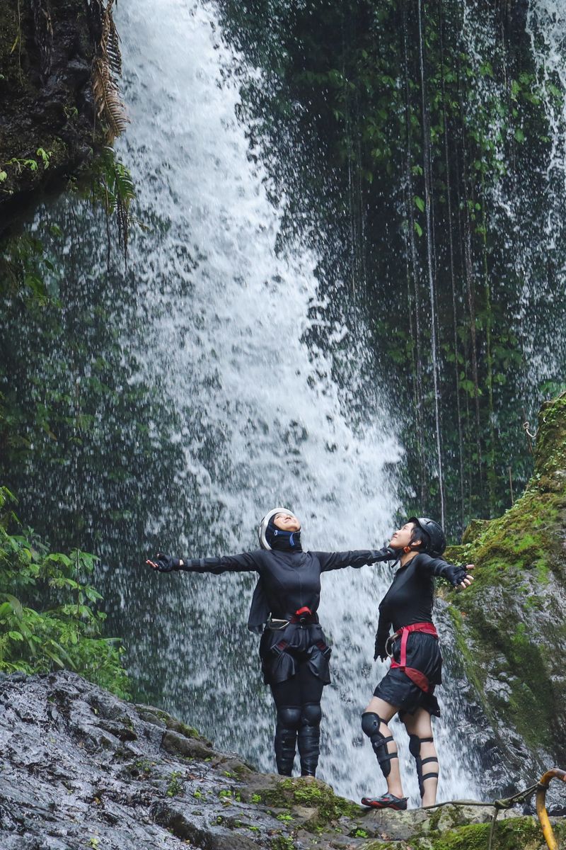 Two Female Adventurers Standing On Mossy Rock With Arms Wide Open, Soaking In The Spray Of Massive Waterfall