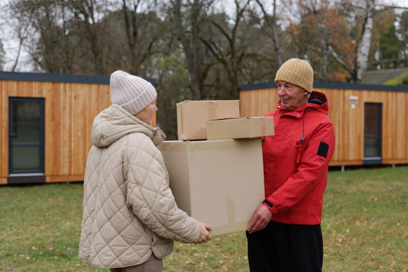 Two individuals are sharing boxes outside near small wooden structures on a cloudy autumn day. One person receives and holds the boxes.