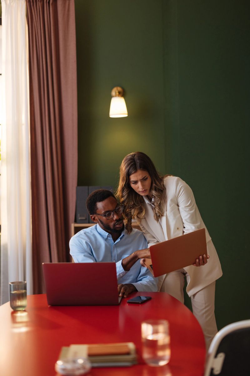 Two professionals collaborate over a laptop and folder in a modern office. The red table, green wall, and warm light create a focused, professional atmosphere for teamwork and discussion.