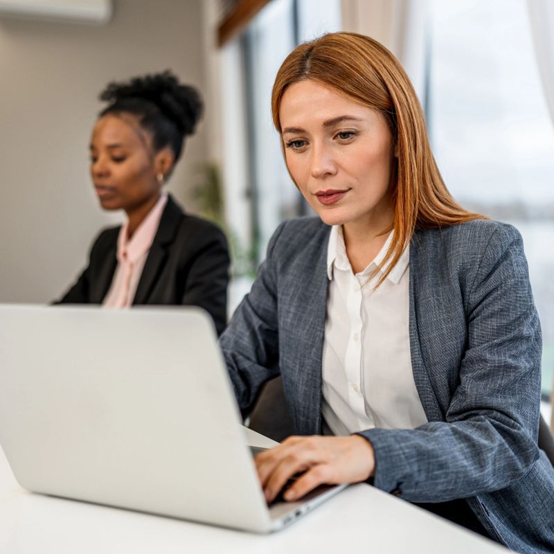 Diverse business women collaborating, focusing on work, and typing on a laptop in a contemporary office environment