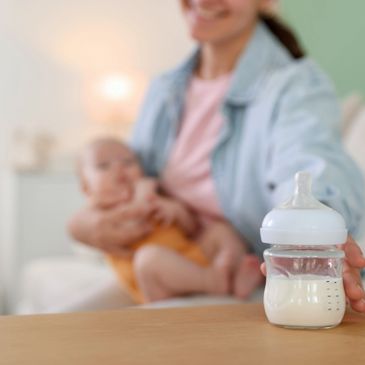 A baby bottle filled with milk on a table, mother holding her baby in the background.