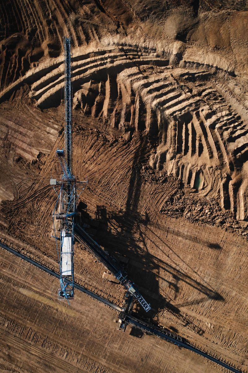 Drone photograph of a massive bucket-wheel excavator operating in a terraced open-pit mine, carving through layers of earth in a radial pattern. The industrial machine casts a long shadow over the textured, brown landscape, highlighting the scale and intensity of surface mining. The image conveys raw power, environmental transformation, and human impact on nature.
