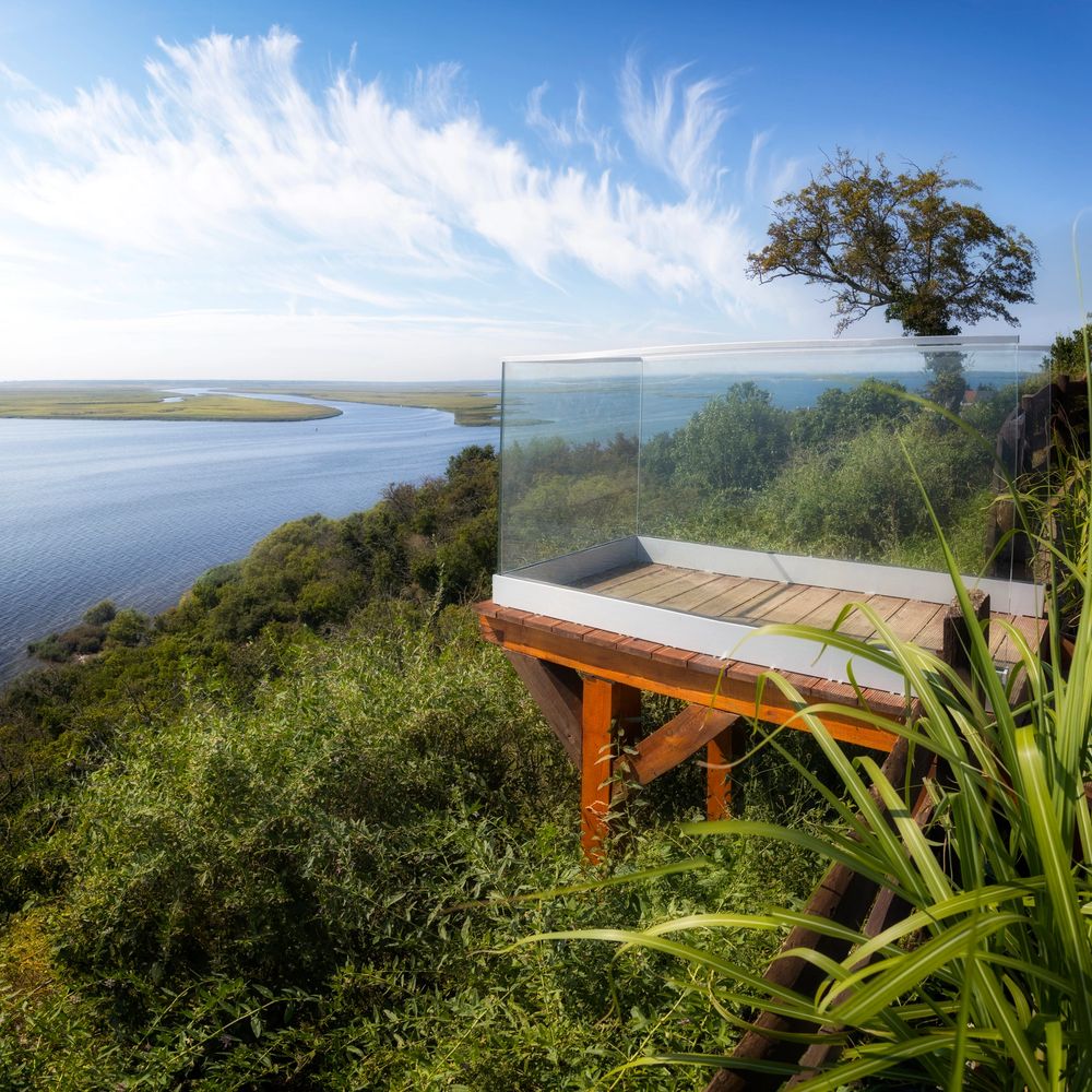 A wooden deck with glass railing overlooking a winding river and green landscape.