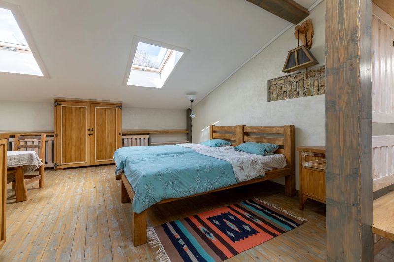 Interior of a warm and bright attic bedroom featuring a natural wood bed with blue linens, ethnic rug on the floor, and skylight windows in a farmhouse style house.