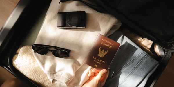 Person packing a suitcase with clothes, camera, sunglasses, and Thailand passport.