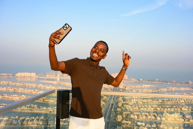 Young adult man happily standing on a rooftop in Dubai, holding up a smartphone to record a selfie video with the iconic Palm Jumeirah and city skyline in the background