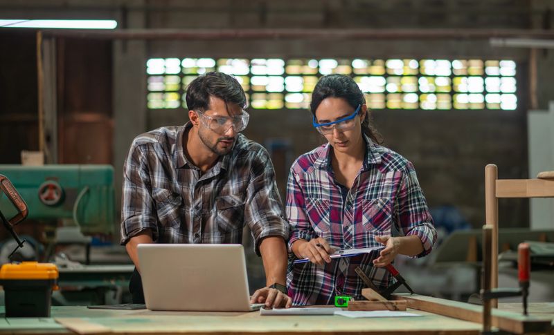 Carpenter workers collaborate reviewing plans and discussing project details clipboard and laptop focus on measurements and construction tasks craftsmanship in a professional industrial setting.