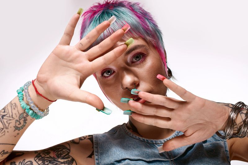 A young woman showcases her artistic style with short, colorful hair and striking nails in a studio.
