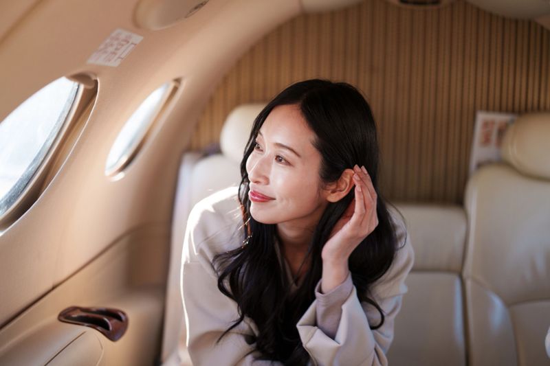 A woman seated inside the cabin of a private jet, looking out the aircraft window during flight. The beige leather interior and oval windows of the jet are visible, highlighting a calm and comfortable travel environment.