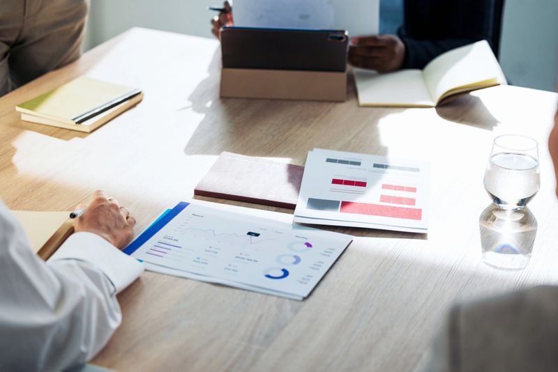 A professional office scene showing a collaborative business meeting with charts, printed reports, notebooks, a tablet, and a glass of water on a clean conference table.