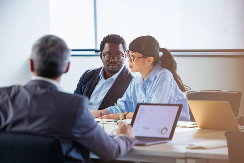 A diverse group of professionals discuss data and ideas around a conference table in a modern office. Laptops and charts suggest collaboration, strategy, and focused teamwork.