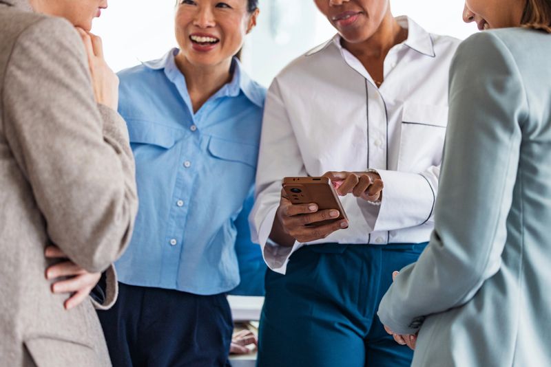 A diverse group of professional women chat and smile as one person shows something on a smartphone. Energetic office vibe with teamwork, collaboration, and positive communication.