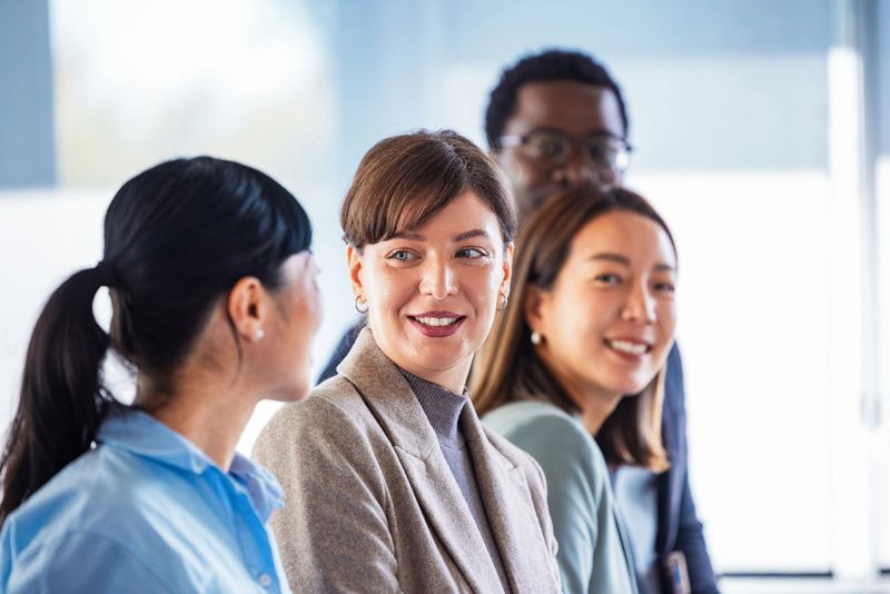 A diverse group of coworkers in a bright office engages in a relaxed discussion. A central smiling woman leads the moment while others listen, signaling teamwork, inclusivity, and a positive business vibe.
