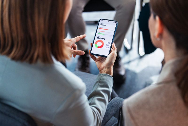 Two women sit together reviewing financial charts on a smartphone, suggesting collaboration, data analysis, and a business discussion in a modern office setting.