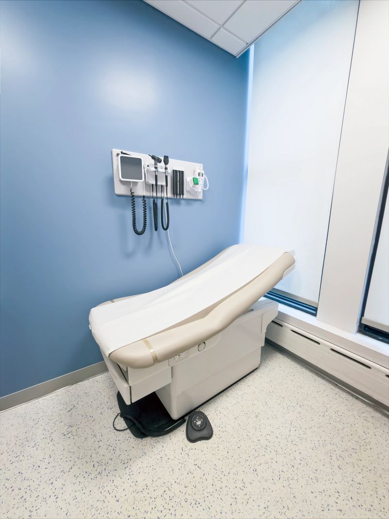 Vertical view of a bright doctor’s examination room featuring a white padded exam table, wall-mounted diagnostic tools, and blue painted walls with a large window letting in natural light. This full frame healthcare interior image is ideal for medical websites, hospital brochures, health insurance marketing, and articles about primary care or clinical visits.