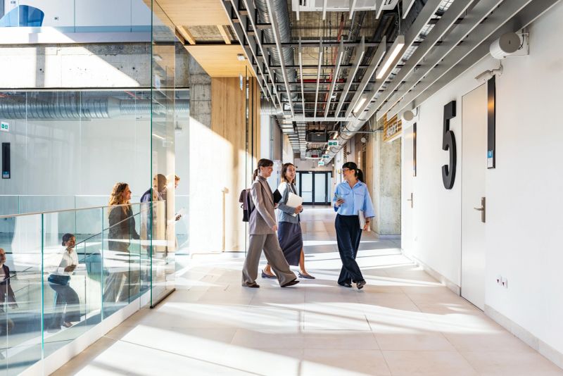 A bright contemporary office hallway where colleagues stroll and chat. Glass railing, exposed ceiling, and large numbers on doors convey a dynamic, professional work environment perfect for corporate, teamwork, and business imagery.