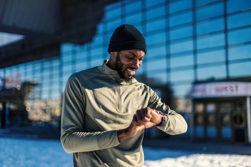 African american man wearing a beanie and athletic top checking his smart watch after an outdoor winter workout, monitoring progress and staying active in cold weather