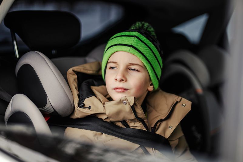 Boy in a green knitted hat and winter jacket gazing thoughtfully out a car window from a booster seat, seatbelt fastened during a calm, safe family road trip journey