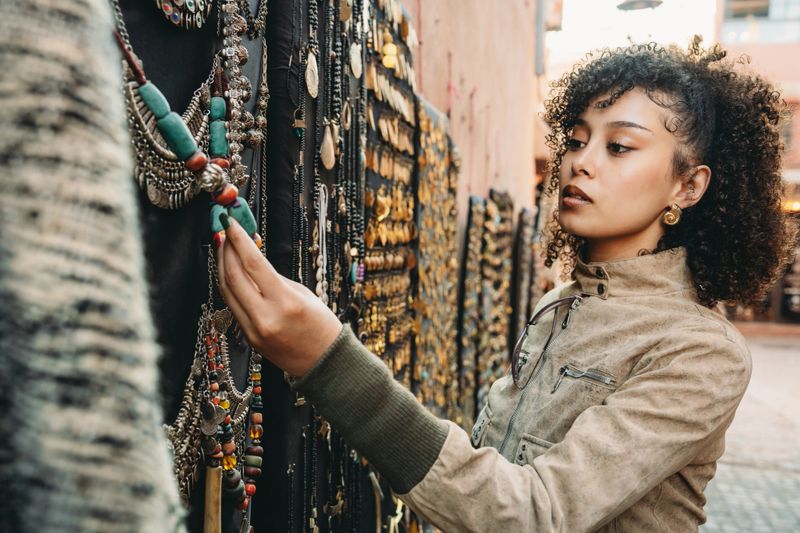 Young Adult woman closely examining a necklace at a colorful street stall filled with ethnic jewelry and handicrafts in a Moroccan market