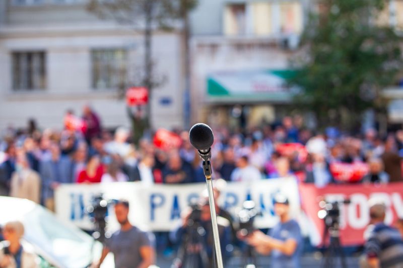 Microphone on podium facing blurred crowd at outdoor political rally, protest or public speech event, leadership and communication concept
