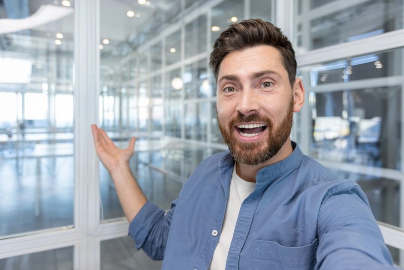 Happy entrepreneur vlogging from a bright modern glass office, smiling and pointing as he introduces the open workspace to his online audience with enthusiasm and confidence