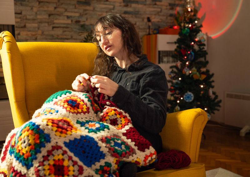 A woman with long brown hair and glasses crochets a granny square blanket while sitting in a big yellow armchair in a cozy living room, while a christmas tree glows in the background and a basket of bright yarn colors sits on the table.