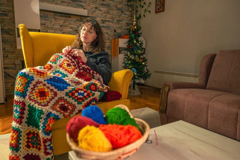 A woman with long brown hair and glasses crochets a granny square blanket while sitting in a big yellow armchair in a cozy living room, while a christmas tree glows in the background and a basket of bright yarn colors sits on the table.