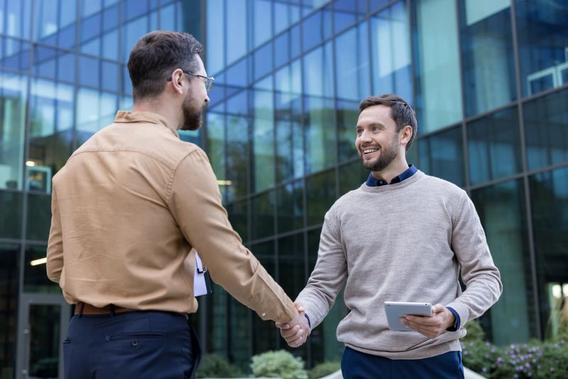 Two smiling men in smart casual attire meeting and shaking hands outside a contemporary glass-fronted office building, completing a successful agreement