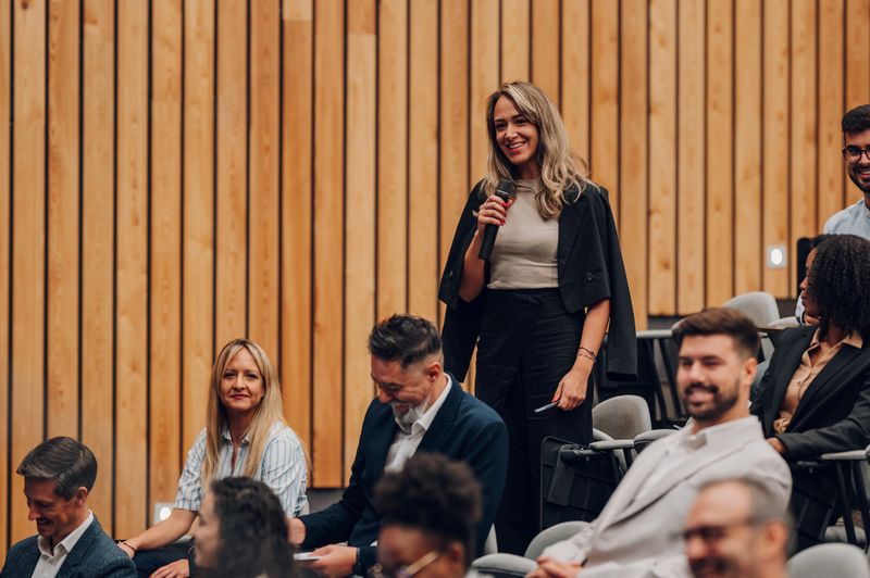 Businesswoman standing in an audience holding a microphone and smiling while asking a question at a corporate event