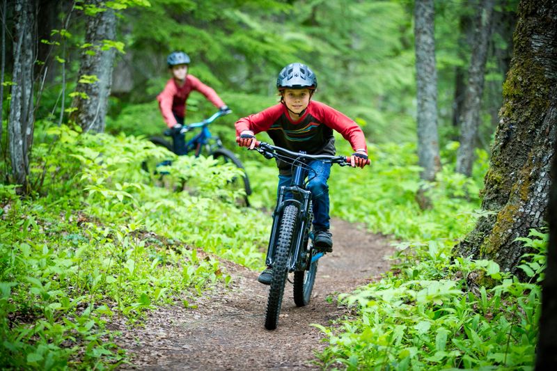 Two nine-year-old boys ride down a smooth, singletrack mountain bike trail near Nelson, British Columbia, Canada, in the summer. They both ride modern full-suspension mountain bikes. They wear cycling jerseys, jeans, bicycle gloves and shoes, and bicycle helmets.