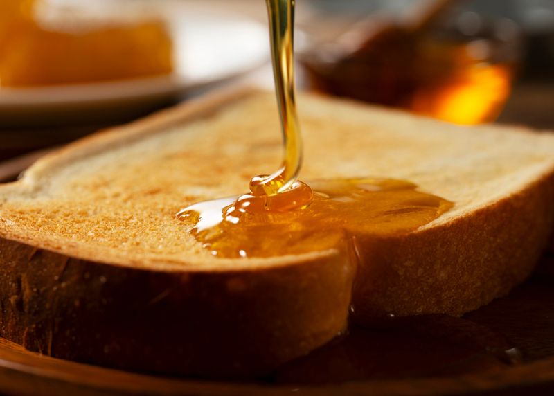 Pouring Fresh Golden Honey onto a Toasted Bread Slice for Breakfast, Delicious Organic Sweetener on Wooden Plate with Copy Space