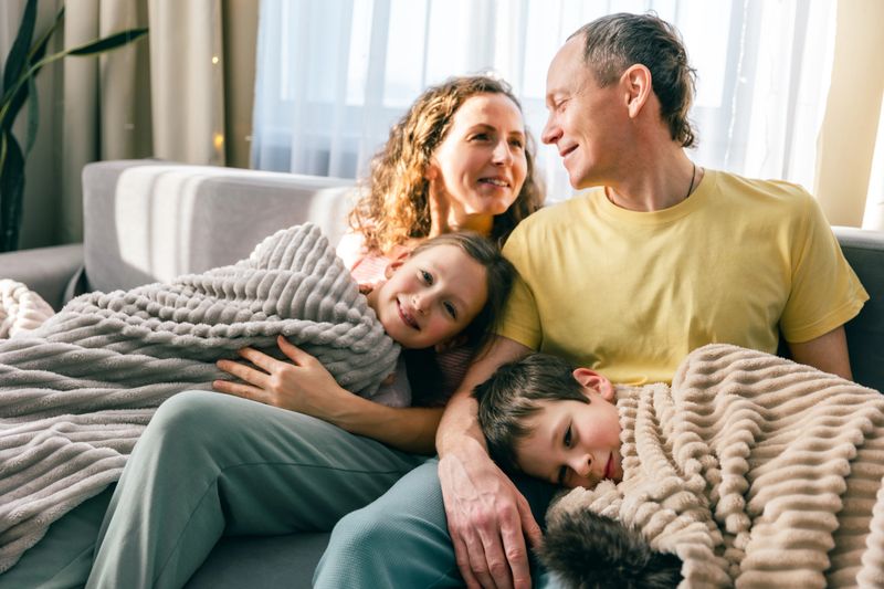 Smiling parents relax on sofa with two children wrapped in cozy blanket at home. Warm family moment, comfort, safety, love and emotional connection, peaceful lifestyle scene with copy space.