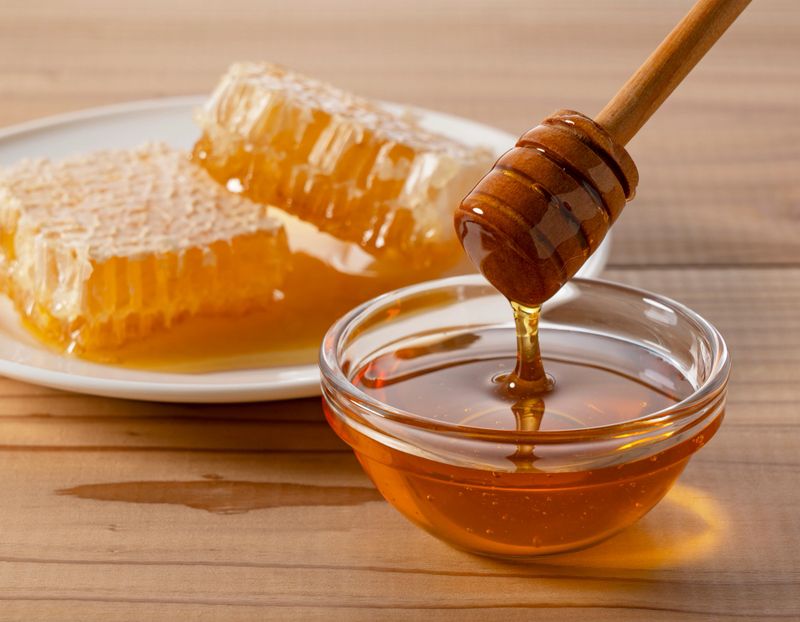 Pure Organic Honey Dripping from Wooden Dipper into Glass Bowl with Honeycomb on Rustic Wood Table