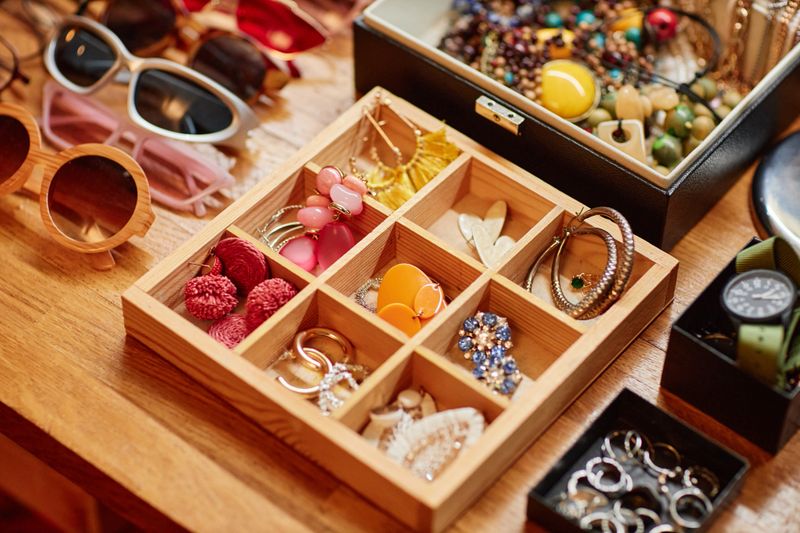 Assorted second hand jewelry and accessories arranged in wooden organizer on table, including earrings, rings, bracelets, sunglasses, watches, showing variety of styles and materials