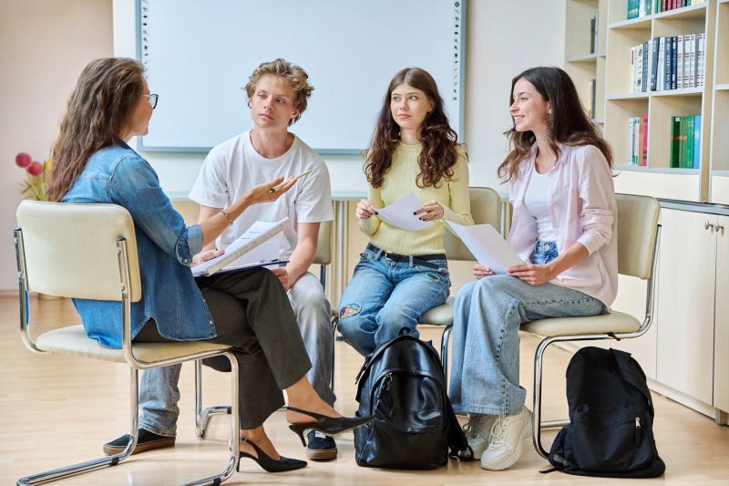 Group teamwork, students teenagers sitting in a circle with a female teacher tutor mentor coach counselor inside a library classroom. Psychology, group mental therapy, education, youth concept