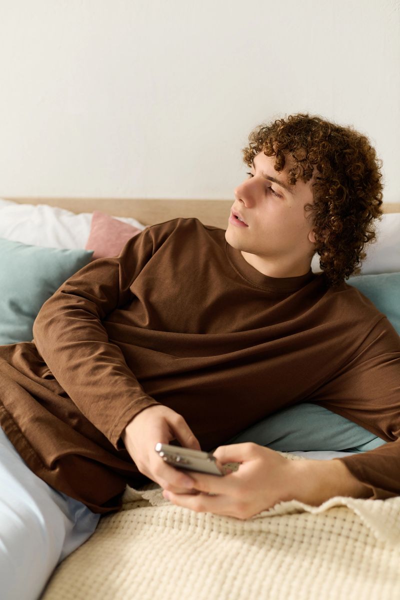 Teenager with curly hair lounges on his bed, focused on his smartphone in a modern apartment.