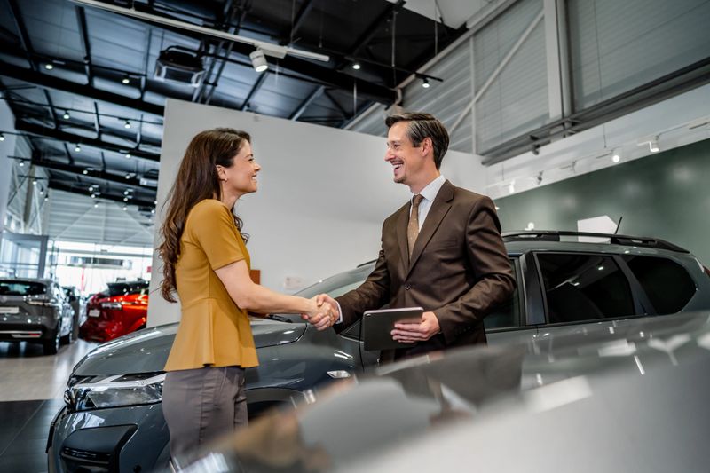 Smiling car salesman and female customer shaking hands in a modern dealership showroom, celebrating a successful purchase