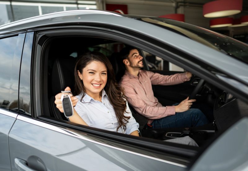 Young woman smiling, proudly holding a car key from the driver's seat of her new vehicle, with her partner beside her